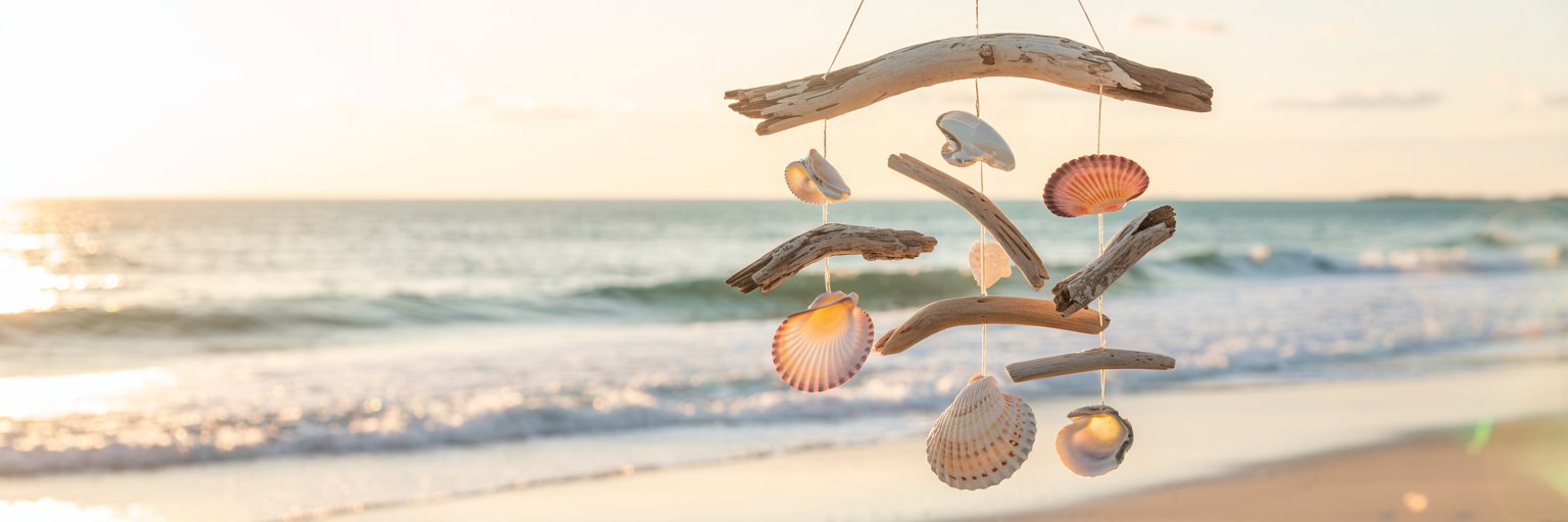 Ein Mobile aus Treibholz und Muscheln hängt im Vordergrund an einem Strand bei Sonnenuntergang, im Hintergrund sind sanfte Meereswellen zu sehen und die Sonne scheint am Horizont.